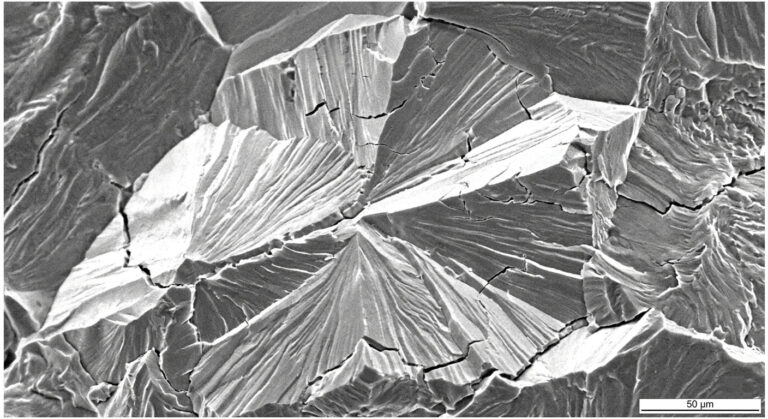High-magnification SEM micrograph showing transgranular cleavage facets and river patterns on a brittle fractured steel surface, indicative of cold-temperature structural failure.